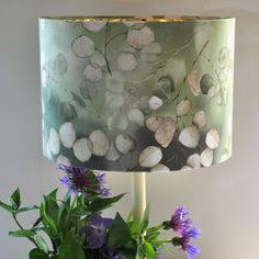 a green lamp shade with purple flowers on it next to a blue vase filled with greenery