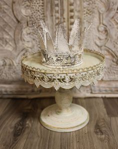 a white cake stand with a crown on it's top sitting on a wooden floor