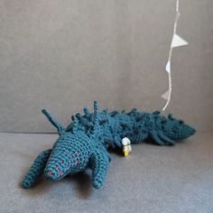 a crocheted blue lizard laying on the ground next to a string with a white ball in it's mouth