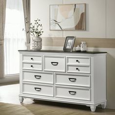 a white dresser with drawers and pictures on the top, in front of a window