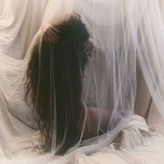 a woman with long hair sitting in front of a white curtain covered bed sheet and holding her head