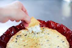 a hand dipping a piece of bread into a casserole in a red dish