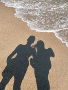 two people standing in the sand next to the ocean with their shadow on the beach