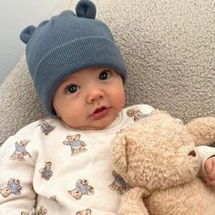 a baby is sitting next to a teddy bear wearing a blue hat and holding a brown teddy bear