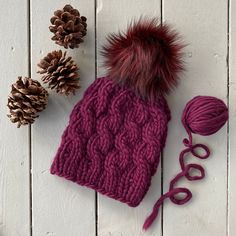 a knitted hat with a pom - pom sits next to some pine cones