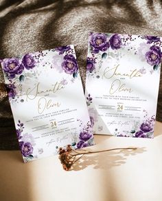 two wedding cards with purple flowers on them sitting on a bed next to each other