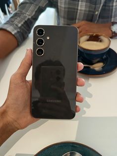 a person holding up a samsung phone in front of a cup of coffee on a table