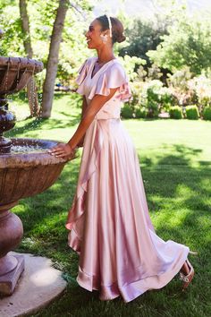 a woman in a long pink dress standing next to a fountain and looking off into the distance