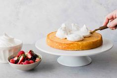 someone is spreading whipped cream on top of a cake with strawberries in a bowl