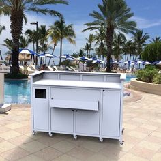 a white cooler sitting next to a swimming pool with palm trees and umbrellas in the background