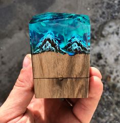 a hand holding a small wooden box with mountains painted on the side and wood in the middle