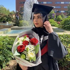 a woman wearing a graduation cap and gown holding a bouquet of flowers in front of a fountain