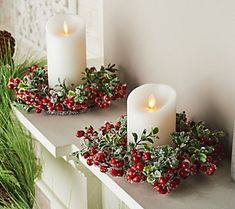 two white candles sitting on top of a mantle next to holly wreaths and pine cones