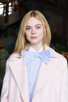 a woman with blonde hair wearing a pink coat and blue bow tie, looking at the camera