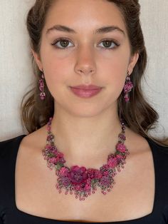 a woman wearing a necklace and earrings with pink flowers on the front, standing in front of a white wall