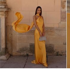 a woman in a long yellow dress standing next to a wall and holding a purse