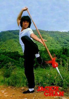 a man holding a wooden stick on top of a dirt road next to a lush green hillside