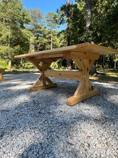 a wooden table sitting on top of a gravel covered ground next to trees and bushes