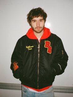 a man wearing a black and red jacket standing in front of a white wall with his hands in his pockets