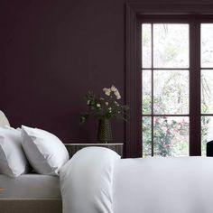 a bed with white sheets and pillows in front of a purple painted wall, next to a window