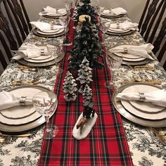 the table is set for christmas dinner with red and black plaid runneres, silver place mats, white plates, and evergreens