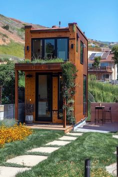 a small wooden house with plants growing on the roof and windows, in front of a hillside