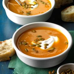 two bowls of carrot soup with bread and pumpkin seeds on the side next to it