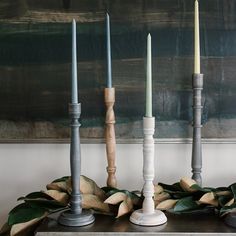 four candles are lined up on a table with greenery and leaves in front of a painting