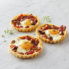 three small tarts with eggs and bacon on them sitting on a marble countertop