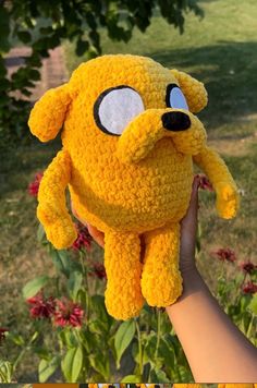 a hand holding a yellow stuffed animal in front of some flowers and trees with eyes