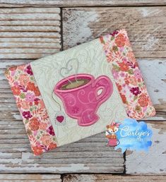 a pink coffee cup on top of a white wooden table next to a piece of fabric