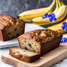 two slices of banana bread on a cutting board next to some bananas and blue flowers