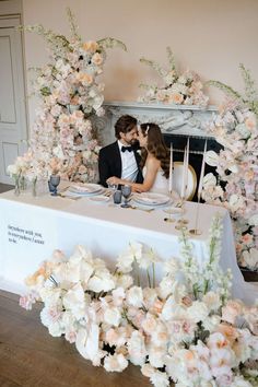 a bride and groom sitting at a table with flowers in front of the fire place