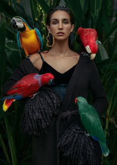 a woman with four parrots on her shoulders in front of plants and foliage,