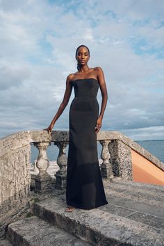 a woman in a black dress standing on some steps by the ocean with her hand on her hip