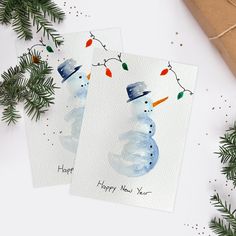 two christmas cards with snowmen on them next to evergreen branches and presents wrapped in brown paper