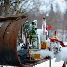 a wooden barrel filled with alcohol bottles on top of a snow covered ground next to a christmas tree