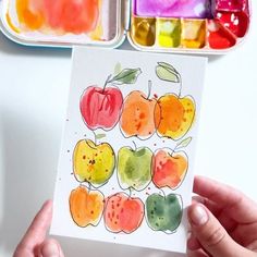 someone holding up a card with watercolors on it in front of two trays of food