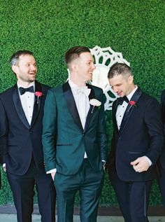 three men in tuxedos standing next to each other near a wall with green grass