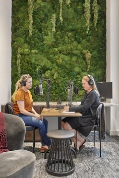 two women sitting at a table talking in front of a wall with plants on it