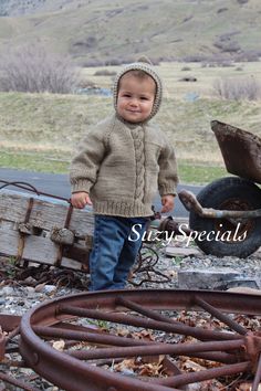 a little boy standing next to an old rusty wheelbarrow and wearing a knitted hat