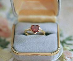 an engagement ring with a heart shaped red stone sits in a box on a floral tablecloth