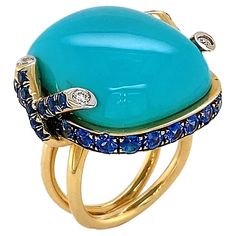 Striking blue of turquoise is the climax of this ring. The polished cabochon is bezel set in 18k yellow gold, which features two inter crossing diamond tipped strands on each side. Sapphires rotate in the bezel setting for a glimmering tranquil accent. Total weights for the gems are 0.14 carats of diamonds, 32.85 carats of turquoise and 3.12 carats of sapphire. The split band complements the setting for a bold finishing touch. Ruby Gold Ring, Teal Jewelry, Yellow Gold Cocktail Ring, Cushion Cut Ring, Gold Cocktail Ring, Contemporary Ring, Blue Sapphire Diamond, Cabochon Ring, Celestial Jewelry