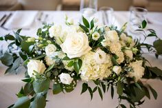 white flowers and greenery are arranged on a table at a wedding reception, with place settings in the background