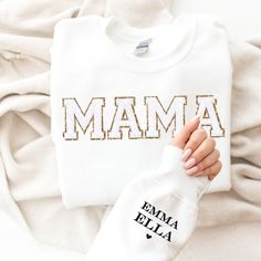 "This cozy crewneck is the perfect gift for a Mama ( or for yourself of course)!  Featuring the word \"Mama\" in bold letters on the front, stylish and cute all year wear. The shirt comes personalized with her kids names on the sleeve. The chenille letter patches are a beautiful white chenille with gold glitter backing, exuding an aura of confidence and vibrancy that's bound to catch everyone's eye. What sets them apart is the sumptuous, fluffy chenille texture that's not only pleasing to the to Winter Name Print Top As Gift, Name Print Tops As Winter Gift, White Tops With Embroidered Graphics For Mother's Day, White Embroidered Tops For Mother's Day, White T-shirt With Letter Embroidery As Gift, Embroidered White Tops For Mother's Day, White Tops With Letter Embroidery For Mother's Day, Cotton Sweatshirt With Lettering For Gift, Embroidered Crew Neck Tops For Gifts