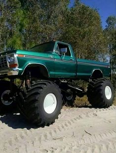 a large green truck with huge tires parked in the sand near some trees and bushes