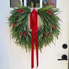 a wreath with red berries and green leaves hanging on a white front door decorated for christmas