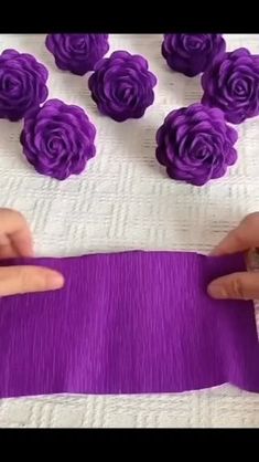 someone is making purple flowers out of yarn on a white tablecloth with pink roses in the background