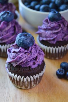 three blueberry cupcakes with purple frosting and fresh blueberries in the background