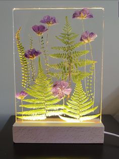 a glass block with purple flowers and ferns in the center on a black table next to a white wall